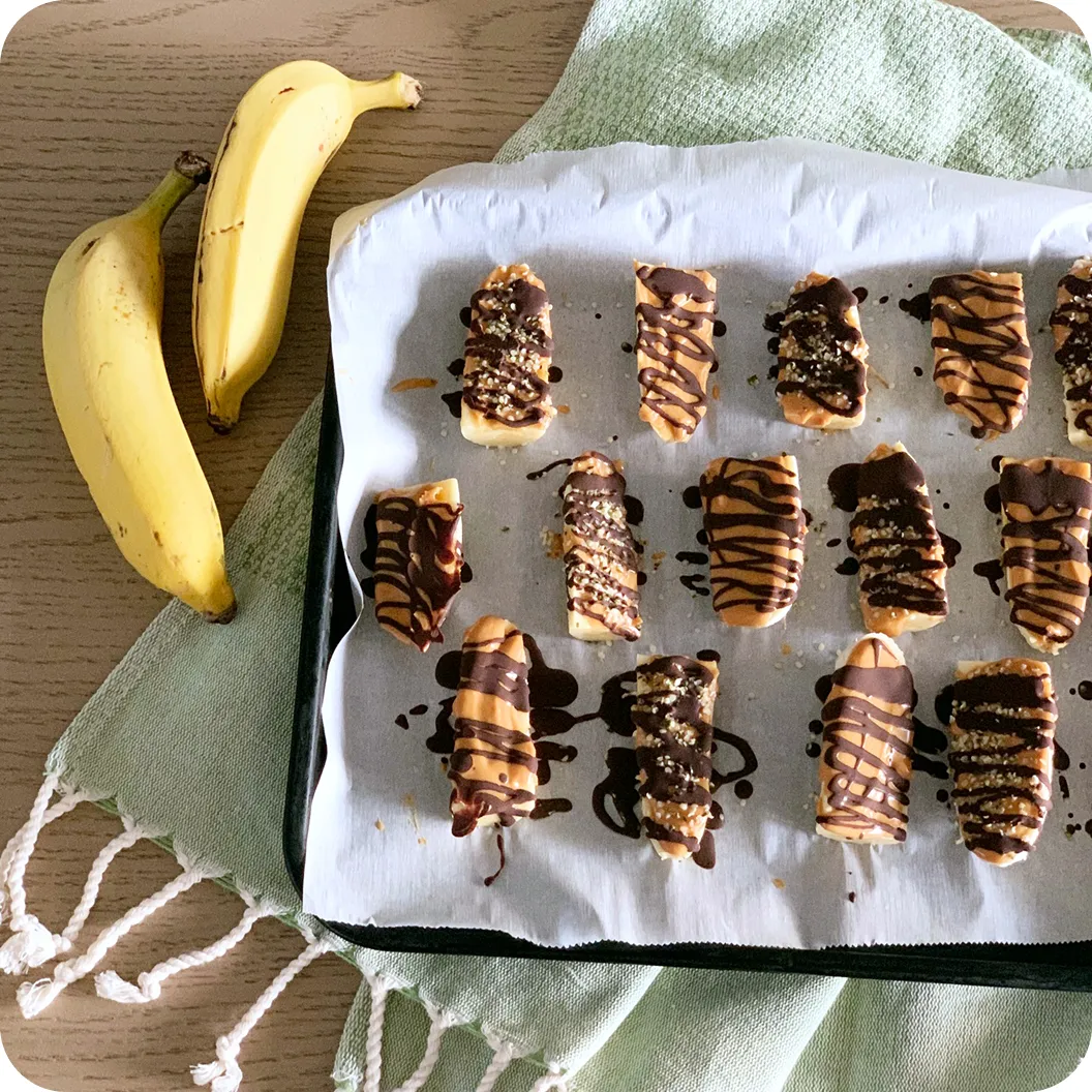 Frozen banana pieces drizzled with peanut butter and dark chocolate on a parchment-lined tray, with fresh bananas beside them