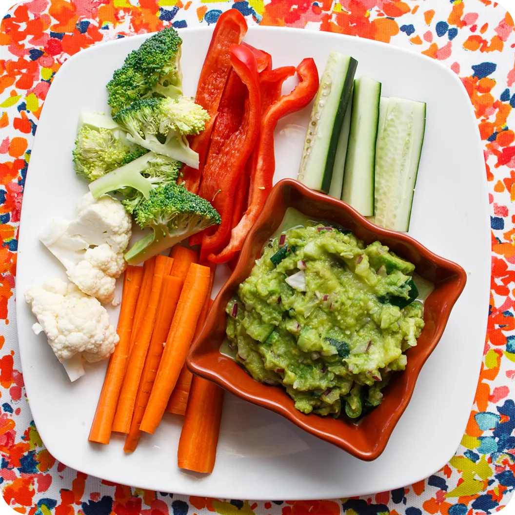 A bowl of chunky guacamole on a plate with carrot sticks, cucumber, red pepper, broccoli and cauliflower