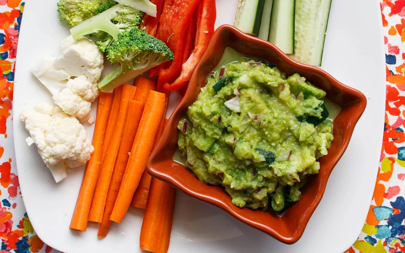 A bowl of chunky guacamole on a plate with carrot sticks, cucumber, red pepper, broccoli and cauliflower