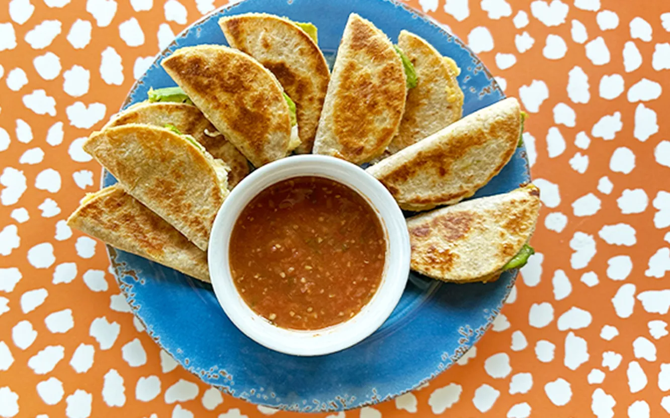 Halved quesadillas fanned around a bowl of salsa on a blue plate, with avocado visible in the filling