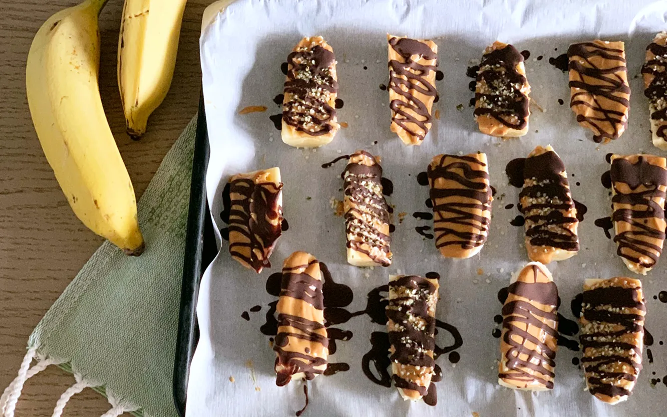 Frozen banana pieces drizzled with peanut butter and dark chocolate on a parchment-lined tray, with fresh bananas beside them