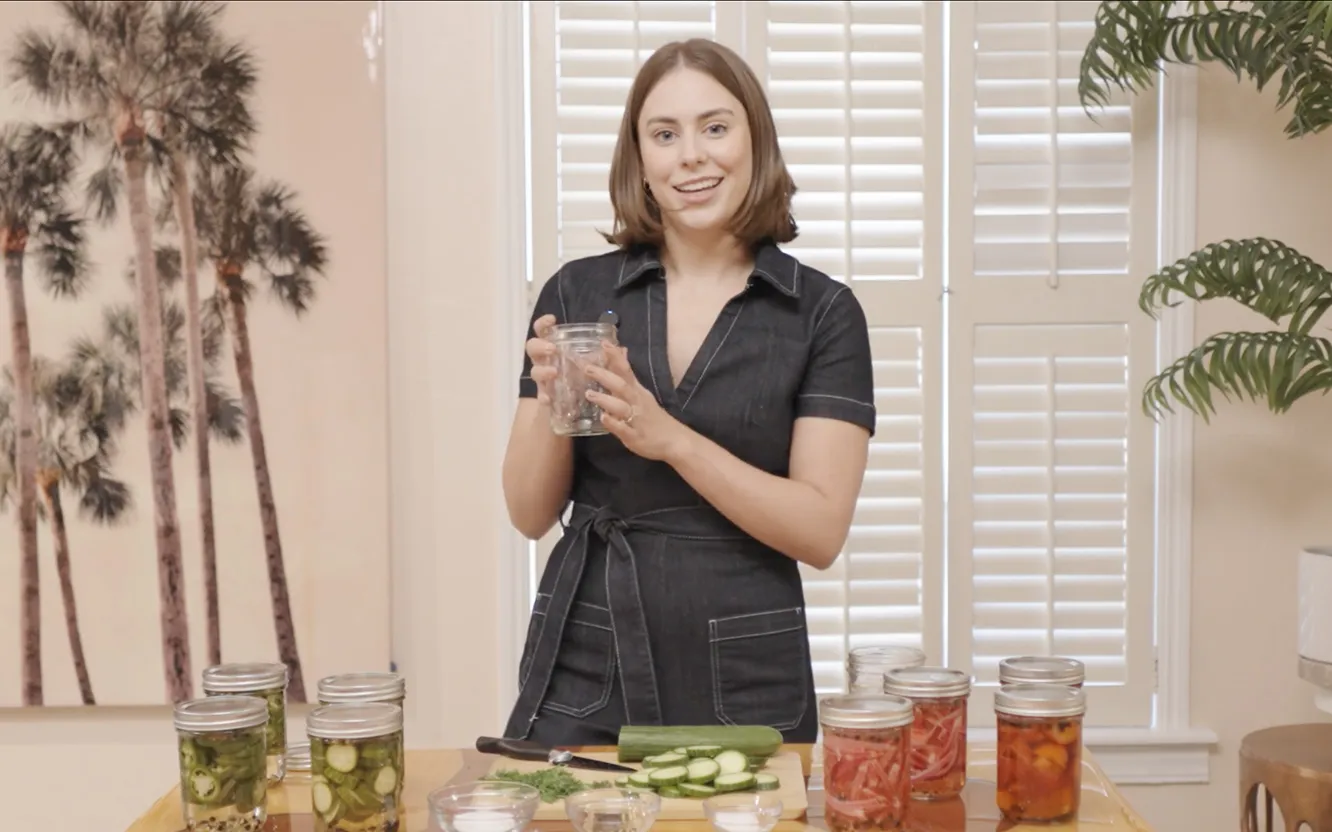 A presenter holds an empty glass jar at a counter lined with jars of pickled okra, cucumber, red onion and peppers.