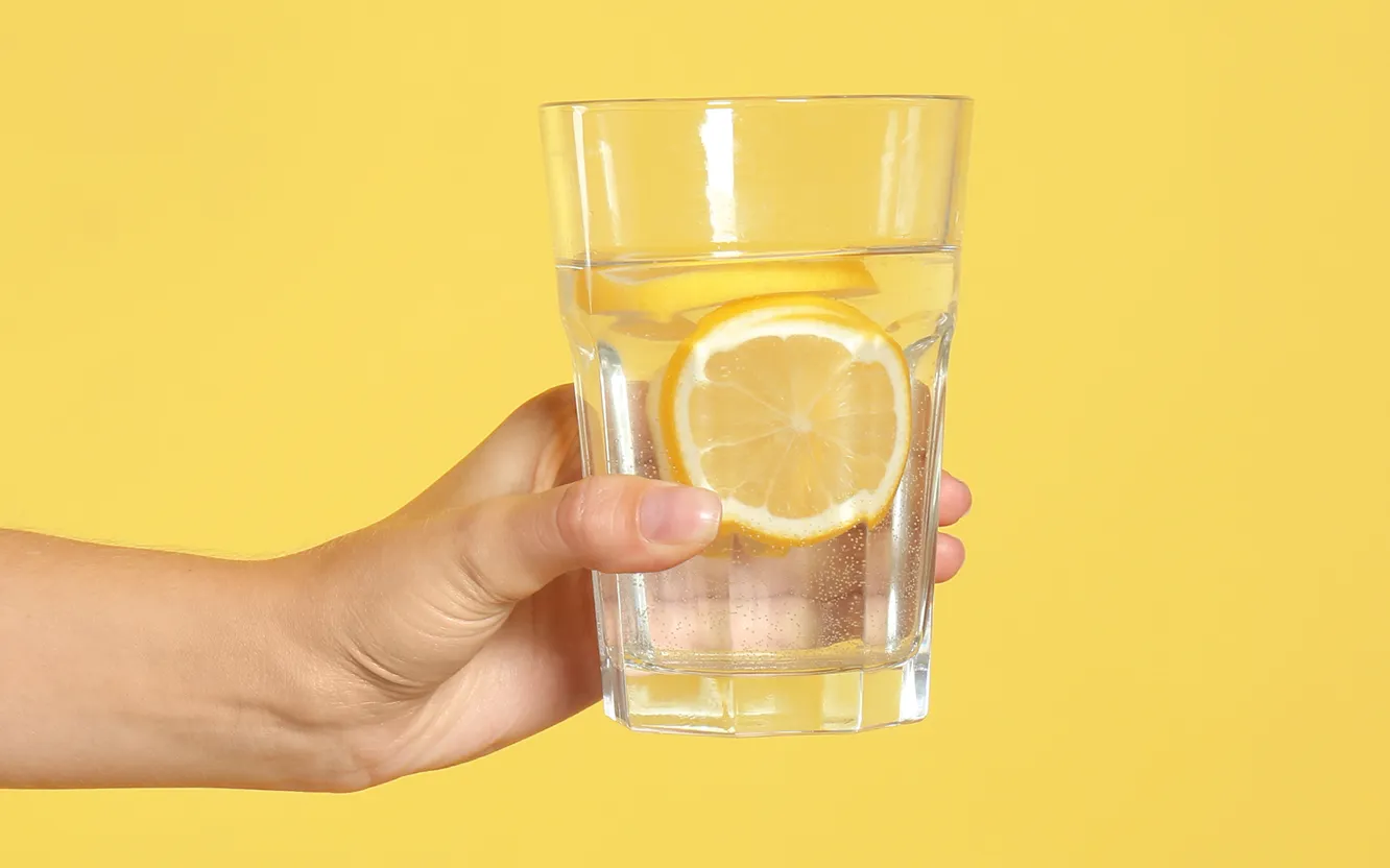 A hand holding a glass of ice water with a lemon slice, against a yellow background